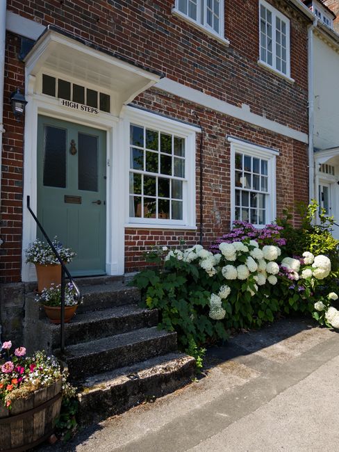 The front of a traditional brick house features a pastel teal front door with a small brass mail slot and a white sign above reading 'High Steers.' White-framed sash windows flank the doorway, with one window behind a lush garden bed filled with large, blooming hydrangeas in white and pale pink, and smaller flowering plants including pink geraniums in a wooden pot to the left of the steps. The entrance is accessed by a short set of weathered stone steps with a black metal handrail, beside which a terracotta pot containing white flowers sits on the top step. The garden borders are well-maintained, with vibrant greenery and flowering bushes extending along a concrete pavement, illuminated by bright natural daylight, highlighting the textures of the brickwork, plants, and garden pots. The scene depicts a neat, charming small front garden emphasizing garden maintenance and landscape care typical of the area, with emphasis on exterior gardening as provided by Gardeners Hounslow.